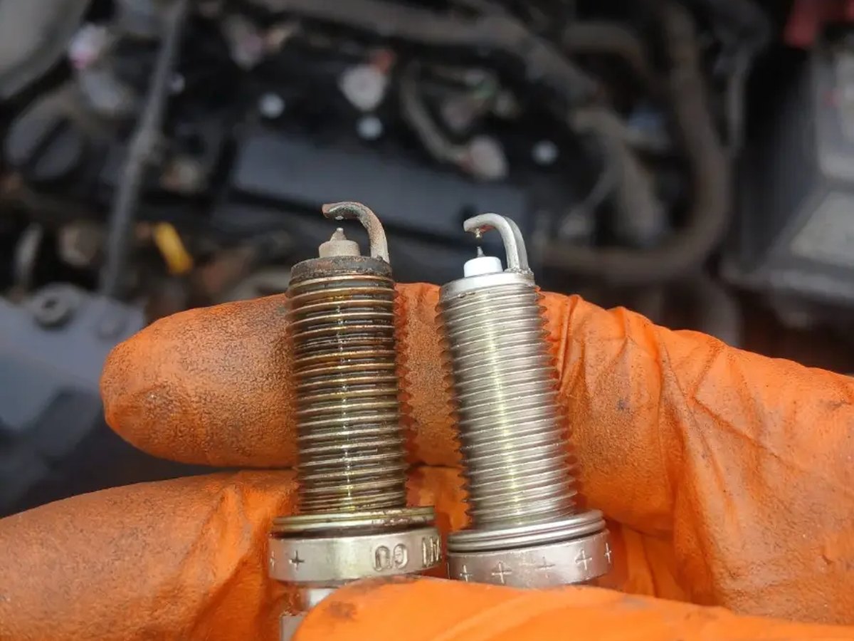 A worn spark plug held next to its new replacement in front of an open engine bay